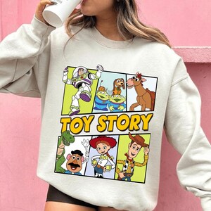 May include: A light gray sweatshirt featuring a colorful graphic of characters from the animated film "Toy Story." The design includes Buzz Lightyear, the aliens, Woody, and other characters, with the words "Toy Story" in yellow.