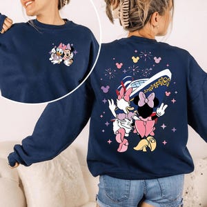 May include: A navy blue sweatshirt with a small embroidered Daisy Duck and Minnie Mouse design on the front. The back features a larger graphic of the characters with a ship, fireworks, and stars. A casual, comfortable style.