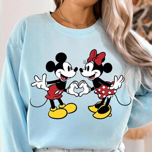 May include: Light blue sweatshirt featuring Mickey Mouse and Minnie Mouse. Mickey is wearing red shorts and yellow shoes, while Minnie is in a red polka dot skirt and a red bow. They are forming a heart with their hands.