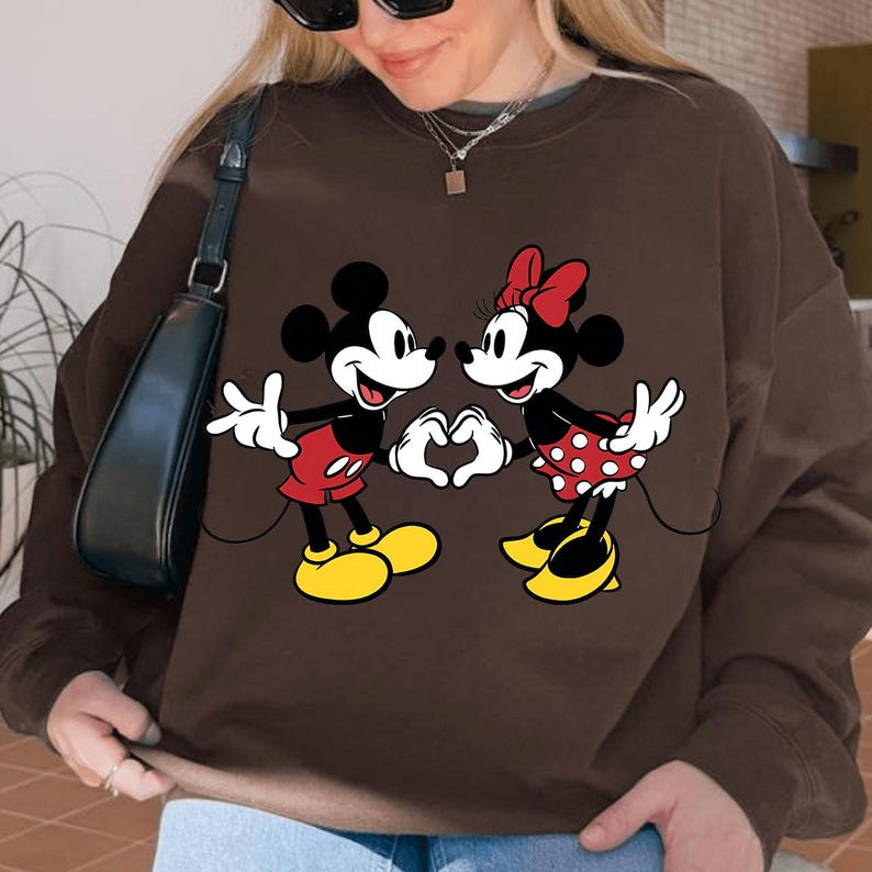 May include: Brown sweatshirt featuring a graphic of Mickey Mouse and Minnie Mouse forming a heart with their hands. Mickey wears red shorts and yellow shoes, while Minnie wears a red polka dot skirt and a red bow.