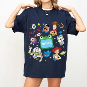 May include: Navy blue t-shirt featuring colorful cartoon characters and the word "NAME" on a green frog-shaped sign. The design includes a rocket ship, stars, and other playful elements.