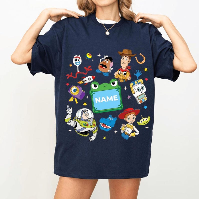 May include: Navy blue t-shirt featuring colorful cartoon characters and the word "NAME" on a green frog-shaped sign. The design includes a rocket ship, stars, and other playful elements.