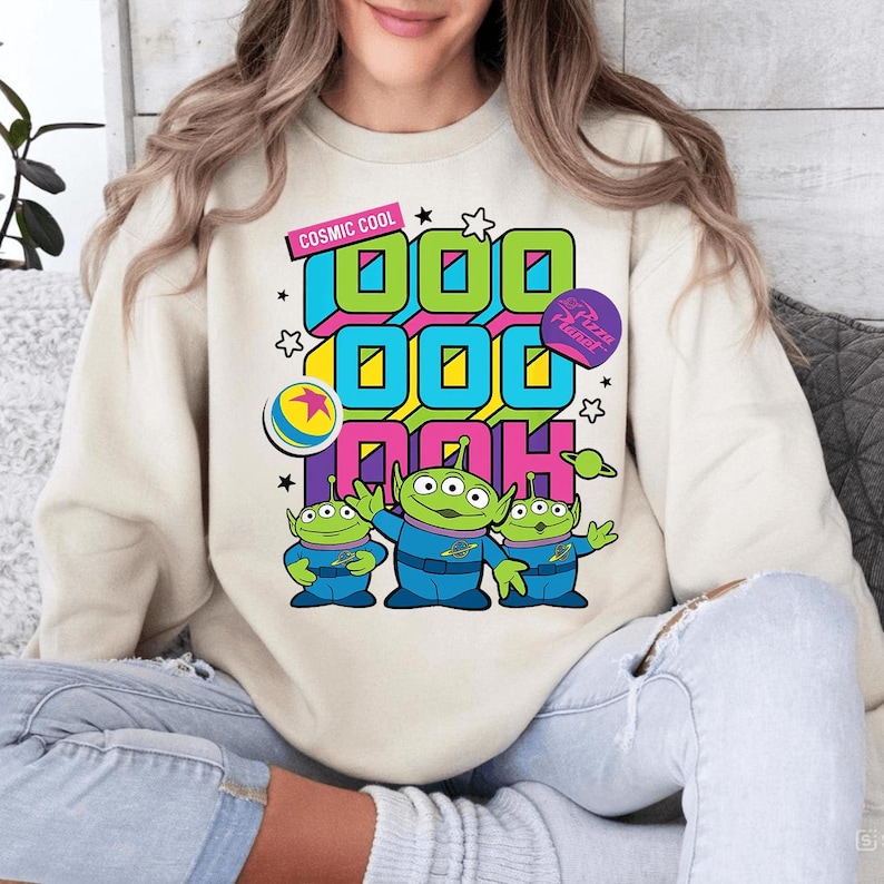 May include: Cream-colored sweatshirt featuring a graphic of three green aliens in blue suits. The design includes the text "COSMIC COOL" and "1000 DOH" in vibrant colors, along with a "Pizza Planet" patch and a ball graphic.
