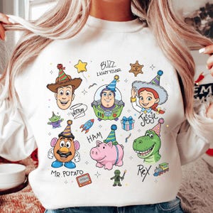 May include: White sweatshirt featuring colorful illustrations of Toy Story characters, including Woody, Buzz Lightyear, Jessie, Mr. Potato Head, Ham, and Rex. The design includes party hats, stars, and the text "Buzz Lightyear".