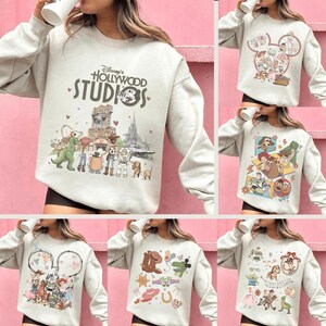 May include: A light gray sweatshirt featuring a Disney's Hollywood Studios graphic. The design includes cartoon characters and iconic park imagery. The sweatshirt has long sleeves and a crew neck.