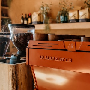 Op de afbeelding: Een koperkleurige La Marzocco espressomachine met de merknaam prominent weergegeven. Een koffiemolen met een transparante bonencontainer staat links. De scène speelt zich af in een café met houten accenten en planken.