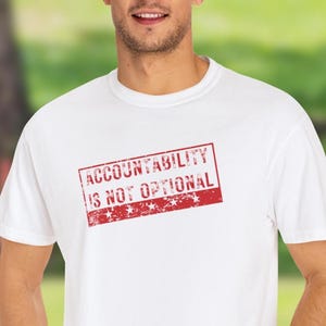 May include: White t-shirt featuring the red text "ACCOUNTABILITY IS NOT OPTIONAL" in a rectangular stamp design. The short-sleeved shirt is made of a soft material. The background is a blurred outdoor scene.