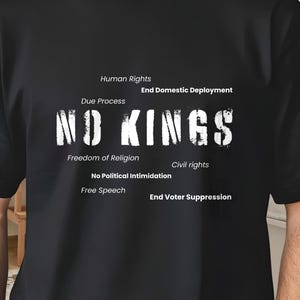 Puede incluir: Camiseta negra con la frase "NO KINGS" en blanco desgastado. Texto adicional incluye "Derechos Humanos", "Debido Proceso", "Libertad de Religión" y otras frases relacionadas. La camiseta es llevada por una persona.