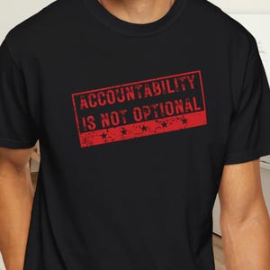 May include: Black crew neck t-shirt featuring the red text "ACCOUNTABILITY IS NOT OPTIONAL" in a rectangular stamp design. The shirt is made of a soft material and is suitable for casual wear.