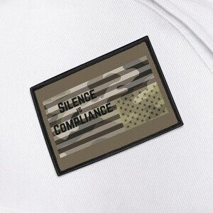 Op de afbeelding: Een witte rugzak met een rechthoekige patch. De patch heeft een camouflage-ontwerp met de tekst "SILENCE IS COMPLIANCE" in zwarte letters. De patch bevat ook een gedeelte met sterren.