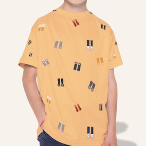 May include: Mustard yellow t-shirt with a repeating pattern of colorful shoe illustrations. The short-sleeved shirt is worn by a young person. The shoes are in various colors, including red, blue, and black.
