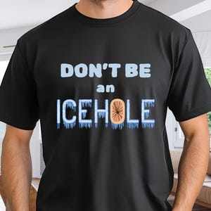 Peut inclure: T-shirt noir avec l'inscription "DON'T BE an ICEHOLE" en bleu clair et orange. Le mot "ICEHOLE" est orné de glaçons. Le t-shirt est porté par une personne.
