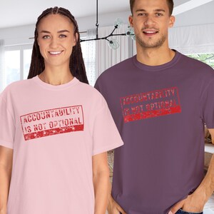 May include: Two people wearing t-shirts. One is light pink, the other is purple. Both shirts have the text "ACCOUNTABILITY IS NOT OPTIONAL" in a red, distressed stamp design.