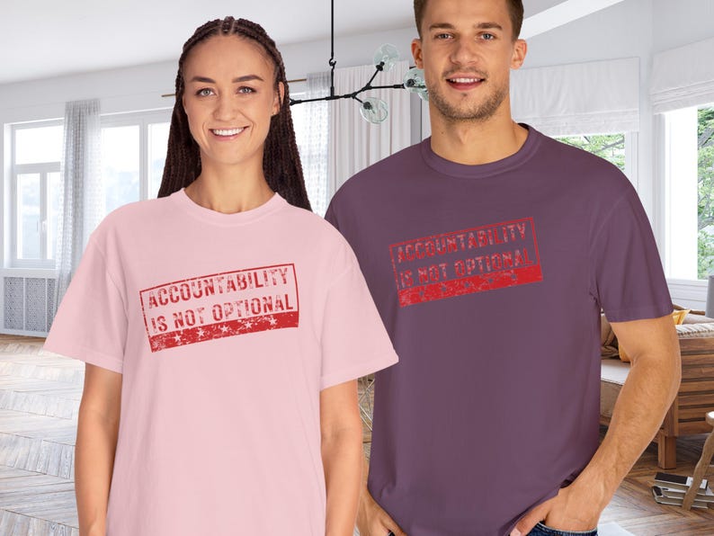 May include: Two people wearing t-shirts. One is light pink, the other is purple. Both shirts have the text "ACCOUNTABILITY IS NOT OPTIONAL" in a red, distressed stamp design.