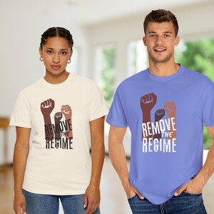 May include: Two people wearing t-shirts with a graphic of three raised fists in shades of brown and tan, with the words "REMOVE THE REGIME" in white. One shirt is cream-colored, the other is light blue.