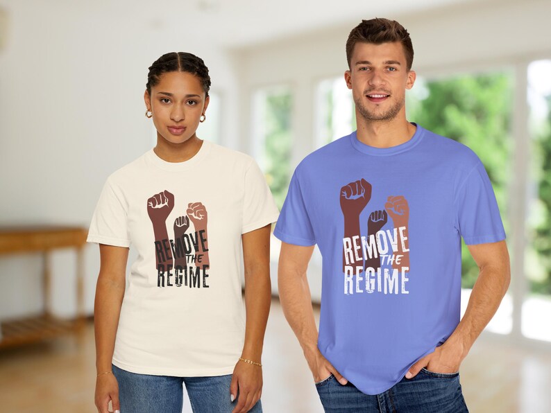 May include: Two people wearing t-shirts with a graphic of three raised fists in shades of brown and tan, with the words "REMOVE THE REGIME" in white. One shirt is cream-colored, the other is light blue.