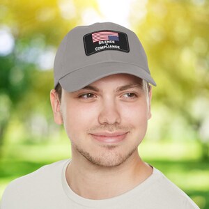 May include: A grey baseball cap with a black patch displaying the American flag and the words "SILENCE" and "COMPLIANCE" in white. The cap is made of a soft material and has a curved peak.