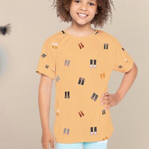 May include: A mustard yellow t-shirt with a pattern of small, stylized boot illustrations in various colors. The shirt is short-sleeved and worn by a young person. The background includes Halloween decorations.