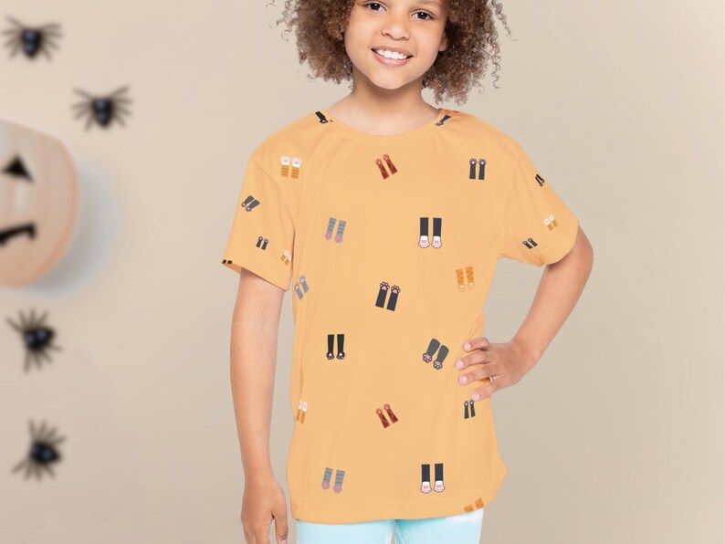 May include: A mustard yellow t-shirt with a pattern of small, stylized boot illustrations in various colors. The shirt is short-sleeved and worn by a young person. The background includes Halloween decorations.