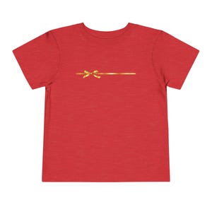 Puede incluir: Camiseta roja para niños pequeños con un diseño de lazo dorado. La cinta es horizontal en el pecho, con un lazo en el centro. La camiseta está hecha de un material suave.