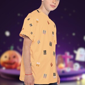 May include: A light orange t-shirt with a pattern of small, colorful shoe illustrations. The short-sleeved shirt is worn by a person. The background is a blurred purple scene with Halloween-themed decorations.