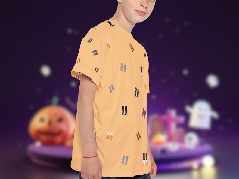 May include: A light orange t-shirt with a pattern of small, colorful shoe illustrations. The short-sleeved shirt is worn by a person. The background is a blurred purple scene with Halloween-themed decorations.