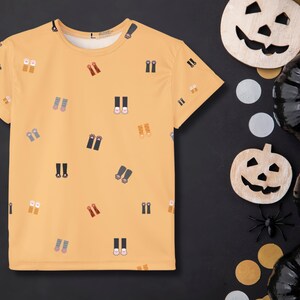May include: A light orange t-shirt with a pattern of various shoes and boots. The shirt is displayed on a black surface with Halloween decorations, including wooden jack-o'-lanterns, black pumpkins, and a plastic spider.