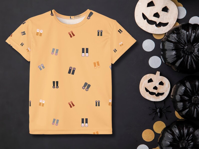 May include: A light orange t-shirt with a pattern of various shoes and boots. The shirt is displayed on a black surface with Halloween decorations, including wooden jack-o'-lanterns, black pumpkins, and a plastic spider.