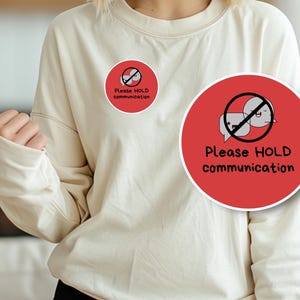 May include: Cream-colored sweatshirt with a round red graphic. The graphic features a speech bubble with a face and a line through it, with the words "Please HOLD communication" in black.