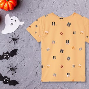 May include: A yellow short-sleeved t-shirt with a pattern of small, colorful binoculars. Halloween decorations, including orange pumpkins, paper bats, spiders, and a ghost, are arranged around the shirt on a gray surface.