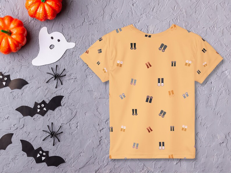 May include: A yellow short-sleeved t-shirt with a pattern of small, colorful binoculars. Halloween decorations, including orange pumpkins, paper bats, spiders, and a ghost, are arranged around the shirt on a gray surface.