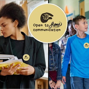 May include: A round, tan button with the words "Open to some communication" and a hand symbol. The button is worn on a black blazer and a blue long-sleeve shirt. The image shows people in a school setting.