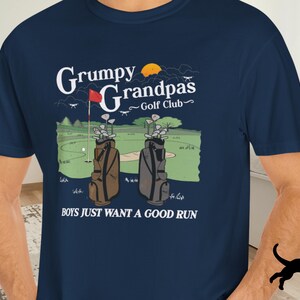 May include: Navy blue t-shirt with the text "Grumpy Grandpas Golf Club" and a golf course graphic. The graphic includes golf bags, a flag, and the phrase "Boys Just Want a Good Run."