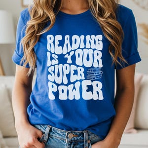Puede incluir: Camiseta azul real con el texto blanco "READING IS YOUR SUPER POWER" en fuente ondulada. El diseño incluye una pila de libros. La camiseta la lleva una persona con vaqueros.