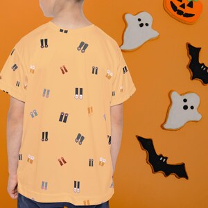 May include: A light orange t-shirt with a pattern of small shoe illustrations. The background is orange with Halloween-themed decorations, including ghost and bat cutouts, and pumpkin cookies.