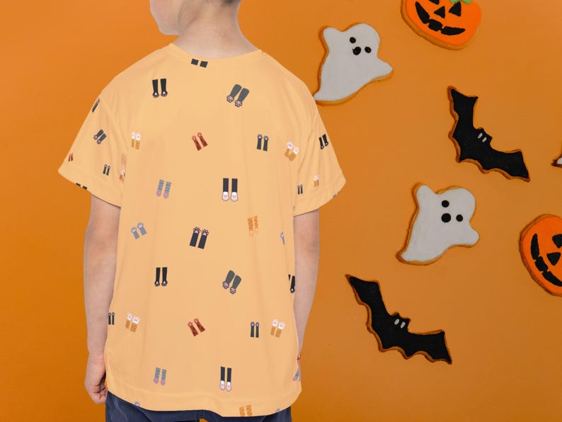 May include: A light orange t-shirt with a pattern of small shoe illustrations. The background is orange with Halloween-themed decorations, including ghost and bat cutouts, and pumpkin cookies.