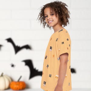 May include: A child wearing a short-sleeved, mustard-colored t-shirt with a repeating pattern of boots. The background features Halloween decorations, including bat silhouettes and pumpkins.