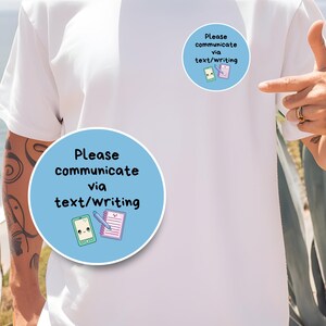 May include: White t-shirt with two light blue circular stickers. Each sticker has the text "Please communicate via text/writing" and illustrations of a smartphone and notepad. The person is pointing at the stickers.