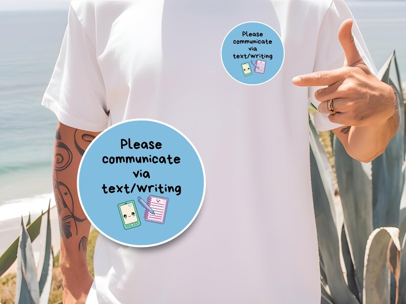 May include: White t-shirt with two light blue circular stickers. Each sticker has the text "Please communicate via text/writing" and illustrations of a smartphone and notepad. The person is pointing at the stickers.