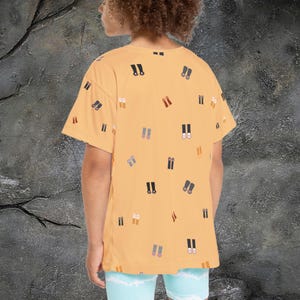 May include: Mustard yellow t-shirt with a pattern of various shoe illustrations in black, brown, and gray. The shirt has short sleeves and a relaxed fit. The person is wearing light blue shorts.