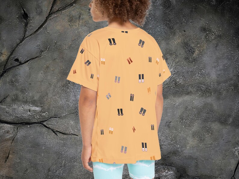 May include: Mustard yellow t-shirt with a pattern of various shoe illustrations in black, brown, and gray. The shirt has short sleeves and a relaxed fit. The person is wearing light blue shorts.