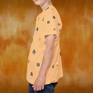 May include: A short-sleeved, peach-colored t-shirt with a repeating pattern of black and white shoe illustrations. The shirt is worn by a person and paired with dark blue pants. The background is a textured orange.