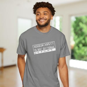 May include: A grey t-shirt with the text "ACCOUNTABILITY IS NOT OPTIONAL" in a white, distressed font. The shirt is a crew neck style and is being worn.
