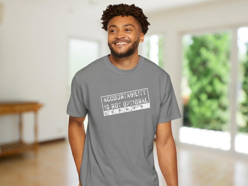 May include: A grey t-shirt with the text "ACCOUNTABILITY IS NOT OPTIONAL" in a white, distressed font. The shirt is a crew neck style and is being worn.