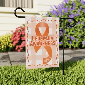 May include: A garden flag featuring a gingham pattern and an orange ribbon design. The flag displays the words "Leukaemia Awareness" with the month "September". The flag is on a black metal stand in a garden.