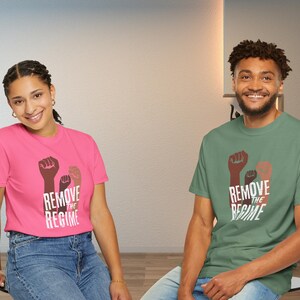 May include: Two people wearing t-shirts with the text "REMOVE THE REGIME" and a graphic of three fists. One shirt is pink, the other is green. Both shirts have short sleeves and are made of a soft material.