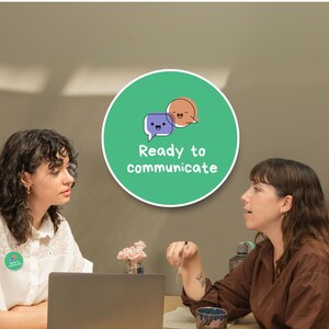 May include: Two women are seated at a table, engaged in conversation. A laptop sits on the table. A green circular sign with the words "Ready to communicate" and speech bubble graphics is visible. One woman wears a white shirt.