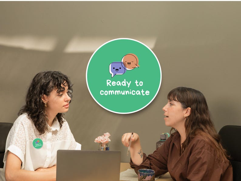 May include: Two women are seated at a table, engaged in conversation. A laptop sits on the table. A green circular sign with the words "Ready to communicate" and speech bubble graphics is visible. One woman wears a white shirt.