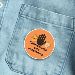 May include: An orange circular button with the text "Communicate only necessities" and illustrations of a hand, fire, and sandwich. The button is pinned to a light blue denim shirt.