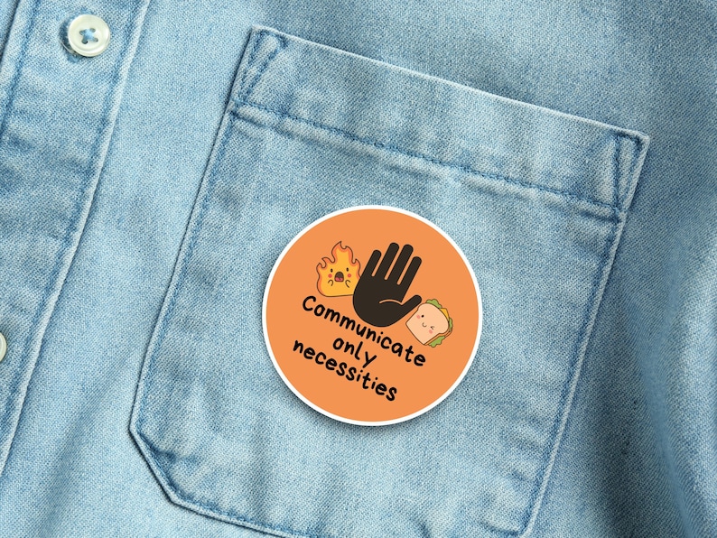 May include: An orange circular button with the text "Communicate only necessities" and illustrations of a hand, fire, and sandwich. The button is pinned to a light blue denim shirt.